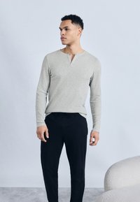 Άνδρας που φοράει ανοιχτό γκρι μπλουζάκι μακρυμάνικο και μαύρο παντελόνι, στέκεται κοιτώντας προς το πλάι μπροστά σε απλό φωτεινό φόντο.