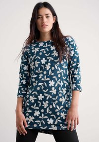 Ženska v temno turkizni tuniki z dolgimi rokavi, okrašeni z belimi cvetličnimi in listnatimi vzorci, stoji pred svetlo enotno ozadje.