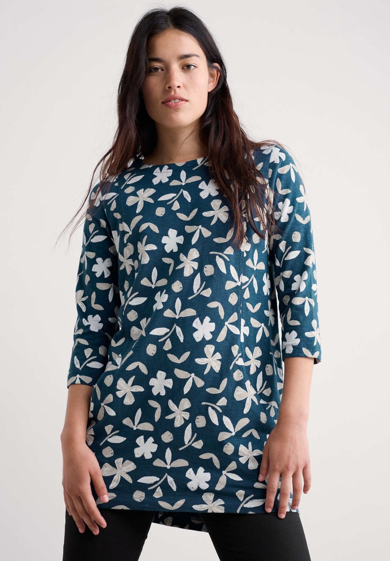 Ženska v temno turkizni tuniki z dolgimi rokavi, okrašeni z belimi cvetličnimi in listnatimi vzorci, stoji pred svetlo enotno ozadje.