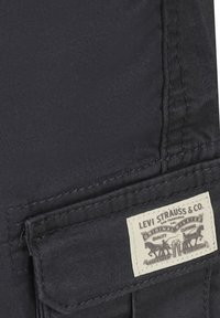 Tessuto nero con cuciture visibili, caratterizzato da un'etichetta rettangolare beige con "Levi Strauss & Co." e un'immagine di cavalli e rivetti.