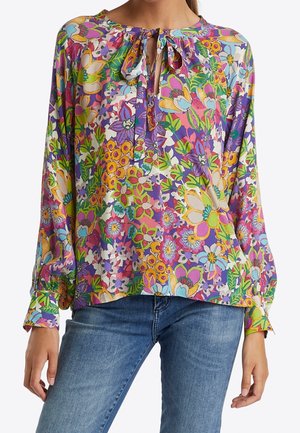 Woman wearing colorful floral blouse with long sleeves and bow tie neck paired with blue jeans, shown from neck to hips.