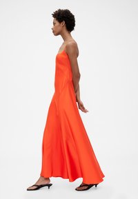 Dlhé oranžové saténové šaty s tenkými ramienkami, voľným strihom a bočnými rázporkami. Modelka má na nohách čierne lodičky, zobrazená z profilu na bielom pozadí.