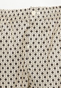 Tessuto beige con motivo geometrico nero che presenta quadrati e forme circolari, inclusi il bottone della vita visibile e le cuciture della tasca.