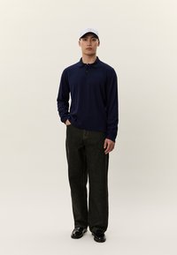 Mann som står, iført marineblå langermet polo, mørke, løstsittende jeans, svarte sko og lys grå baseballcaps, mot en ensfarget lys bakgrunn.