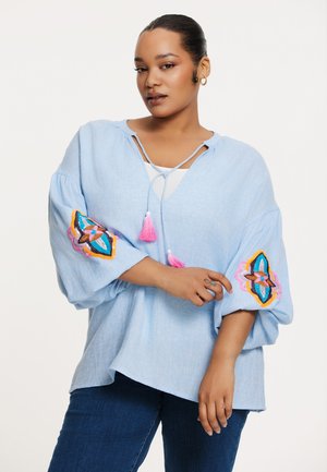 Helles Blau, locker sitzende Bluse mit langen, puffigen Ärmeln. Verziert mit bunten gestickten Akzenten und einem gebundenen Ausschnitt mit Quasten.
