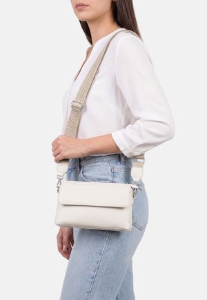 Donna con camicetta bianca e jeans azzurri chiari porta una piccola borsa a tracolla bianca con una larga tracolla beige regolabile.