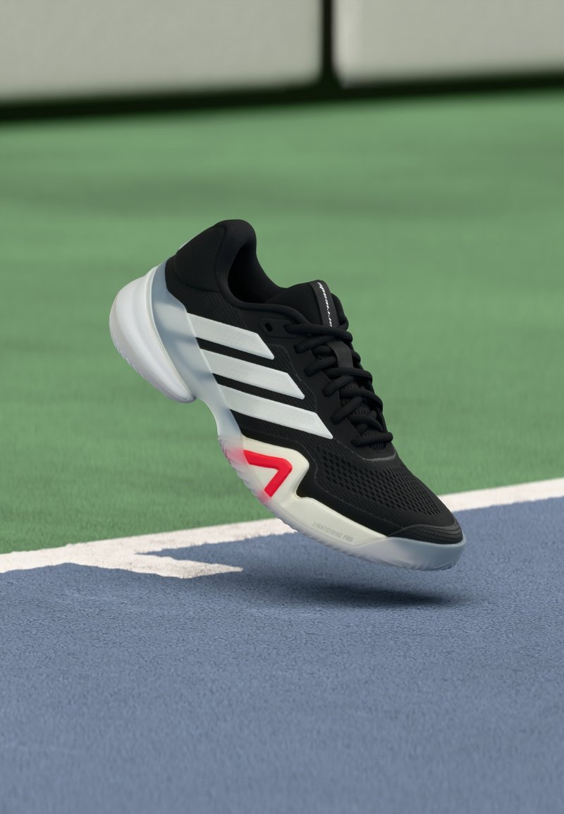 Μαύρο αθλητικό παπούτσι με λευκές ρίγες, υφασμένο μαύρο επάνω μέρος, μαξιλαρωμένη σόλα και κόκκινη λεπτομέρεια στη φτέρνα. Ελαφρύ σχέδιο για απόδοση.