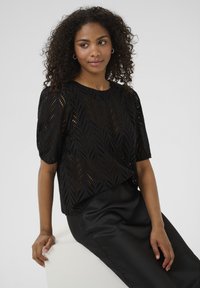 Haut noir à manches courtes avec un design en maille ouverte à motifs de feuilles. Porté sur une jupe noire élégante, mettant en valeur une finition en tissu texturé.