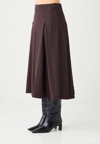 Brauner plissierter Midirock mit hohem Bund aus glattem Stoff, kombiniert mit eleganten schwarzen kniehohen Stiefeln mit spitzer Spitze.