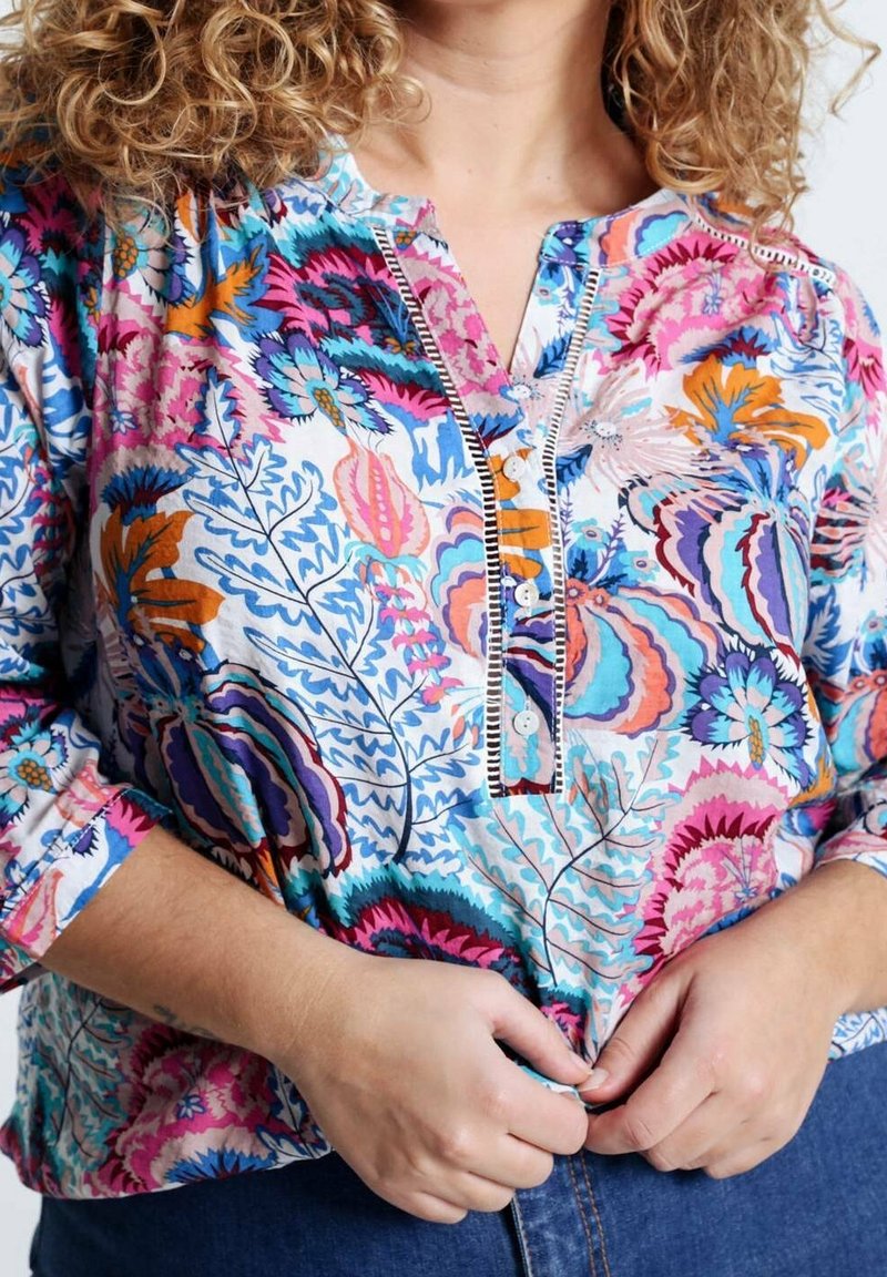 Blouse colorée et à motifs avec des manches longues, patte de boutonnage, ornée de motifs floraux en rose, bleu et orange sur un fond blanc.