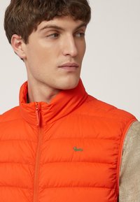 Chaleco acolchado naranja con cuello alto, presenta un zipper vertical y un pequeño logo verde en el pecho. Ligero, con textura acolchada.