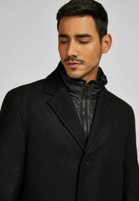 Cappotto nero con esterno in lana testurizzata e un interno trapuntato con zip. Il design include un collo a revers e una chiusura con bottone singolo.