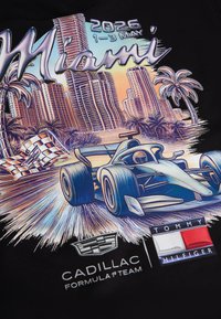 Αυτοκίνητο αγώνα Formula 1 περνά με ταχύτητα τη σημαία με καρό μοτίβο, με το ορίζοντα του Μαϊάμι, φοίνικες και την ημερομηνία της εκδήλωσης 1–3 Μαΐου 2026, λογότυπα Cadillac και Tommy Hilfiger.