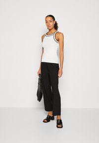 Witte tanktop met zwarte rand en mouwloos ontwerp, gecombineerd met zwarte wijde broek en zwarte sandalen met hakken; houdt een zwarte tas vast.