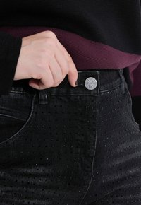Jeans in denim nero con una superficie texturizzata caratterizzata da piccole perforazioni. Bottone argento con dettagli "est. 1987". Maglietta scura indossata sopra la vita.