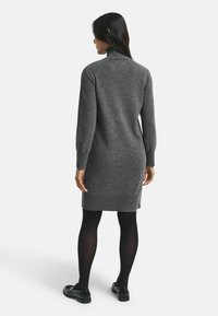 Abito grigio lavorato a maglia con colletto alto, maniche lunghe, polsini e orlo a coste, indossato con collant neri e scarpe, visto da dietro.
