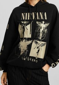 Sudadera negra con capucha que presenta un gráfico dorado de Nirvana con ilustraciones de ángeles, capucha con cordón y un corte holgado. Pantalones negros a rayas usados debajo.