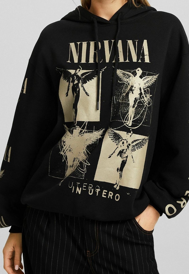 Sudadera negra con capucha que presenta un gráfico dorado de Nirvana con ilustraciones de ángeles, capucha con cordón y un corte holgado. Pantalones negros a rayas usados debajo.