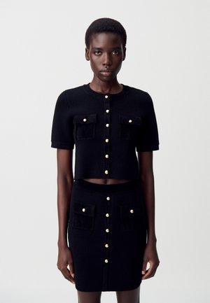 Femme portant un cardigan noir à manches courtes avec boutons et une jupe taille haute assortie, ornée de boutons dorés et de poches avant, se tenant devant un fond blanc.