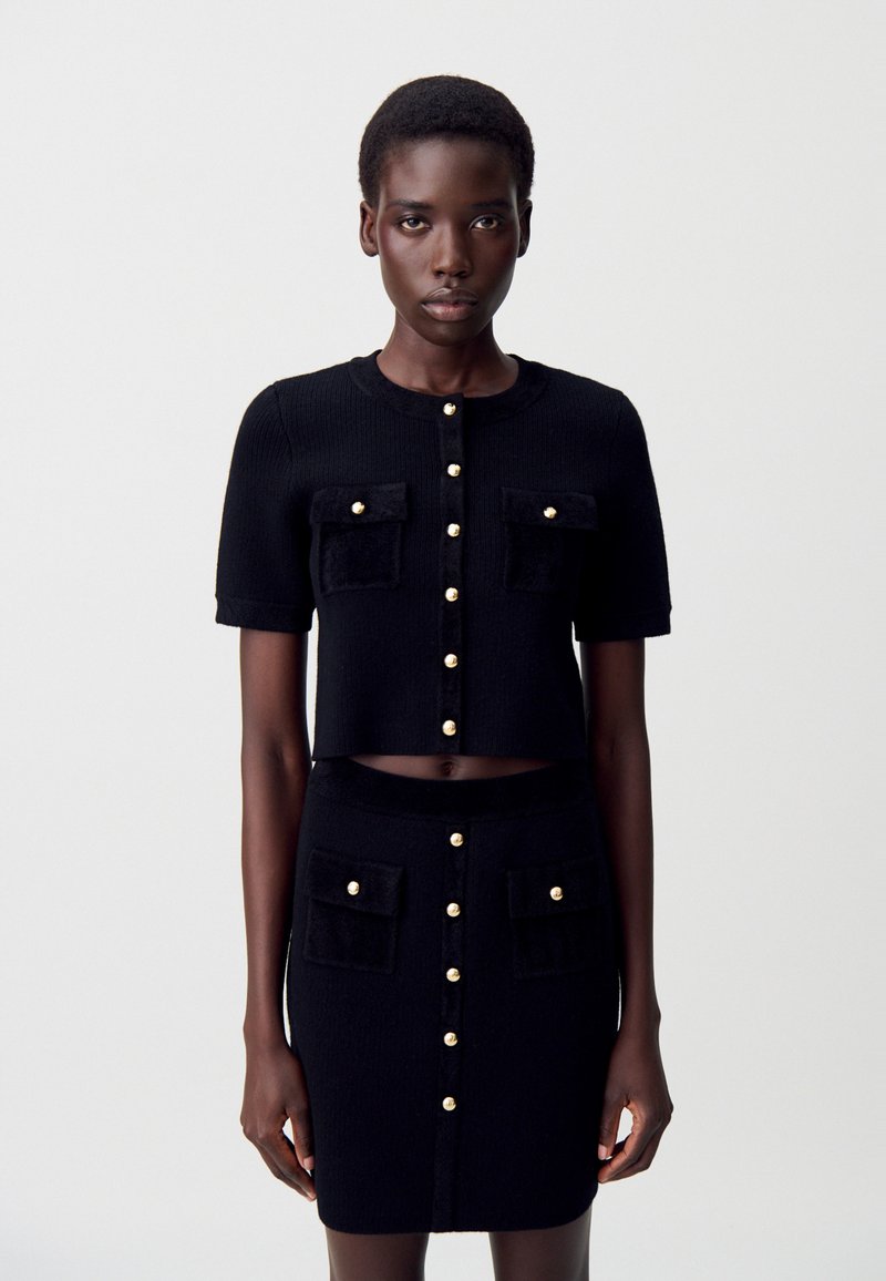 Femme portant un cardigan noir à manches courtes avec boutons et une jupe taille haute assortie, ornée de boutons dorés et de poches avant, se tenant devant un fond blanc.