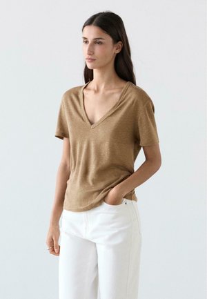 Donna con capelli lunghi e scuri, indossa una maglietta marrone a collo a V e maniche corte e pantaloni bianchi, in piedi con una mano in tasca su uno sfondo neutro.