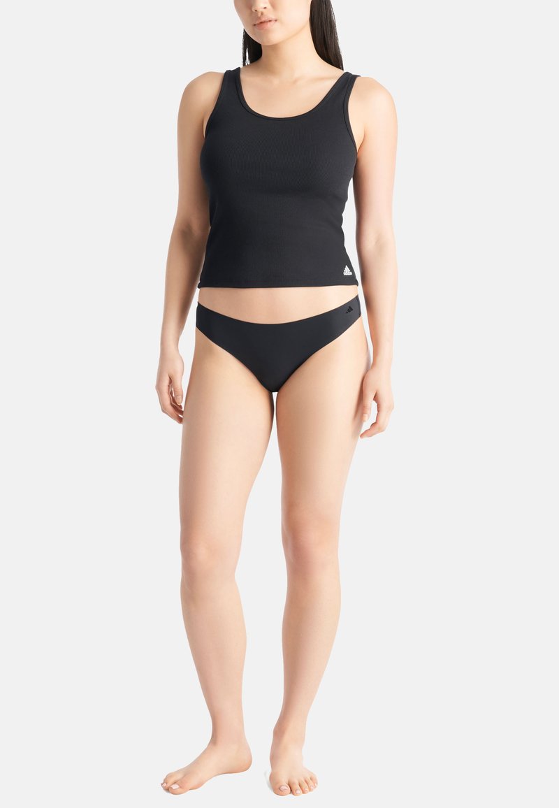 Vrouw in een zwarte geribbelde tanktop en zwarte bikini-achtige onderbroek staat blootvoet tegen een witte achtergrond.