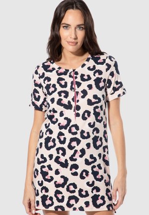 Frau trägt ein kurzärmeliges, pinkes Leopardenmusterkleid mit schwarzen und hellrosa Flecken, steht vor einem schlichten hellgrauen Hintergrund.