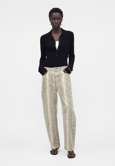 Γυναίκα φορούν μαύρο τοπ με μακριά μανίκια, μπεζ φαρδιά παντελόνια με σχέδιο φίδι, σκουλαρίκια κρίκοι και μαύρα σανδάλια, στέκεται με τα χέρια στις τσέπες.