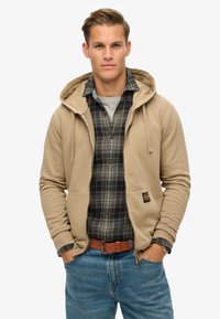Sudadera con cremallera beige con capucha, que cuenta con un bolsillo frontal, llevada sobre una camisa de franela a cuadros. Unos vaqueros azules y un cinturón marrón completan el conjunto.