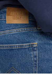 Denim jeans featuring a leather patch labeled "AMERICA TODAY," with visible stitching and a textured blue fabric.