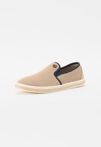 Chaussure beige à enfiler avec un dessus en maille, accents navy et semelle texturée. Présente un bout arrondi et un détail de marque près de l'ouverture.