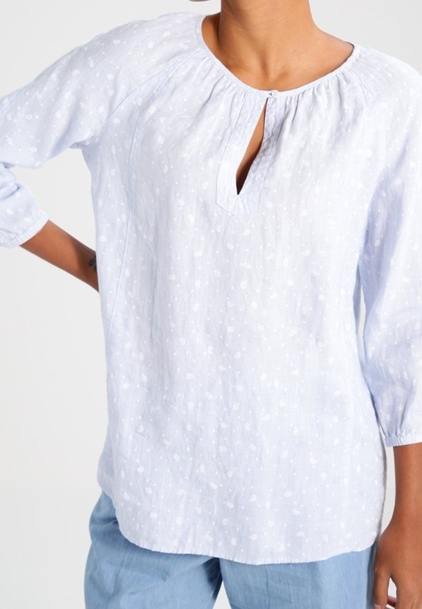 Femme portant un chemisier bleu clair avec un motif floral blanc subtil et des manches trois-quarts, associé à un pantalon bleu clair.