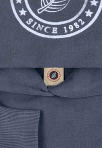 Sweat à capuche en tissu gris foncé avec détails côtelés. Présente un écusson avec le texte "SWEET PANTS SINCE 1982" et un motif de feuille.