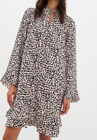 Mekko, jossa on leopardikuosi, kevyeistä kankaista valmistettu, sidottava kaula-aukko, pitkät hihat ja kerroksellinen röyhelöhelma. Värimaailma: beige ja musta.