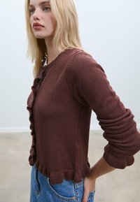 Cardigan marrone lavorato a maglia con un frontale e polsini arricciati, caratterizzato da un collo rotondo e maniche lunghe, abbinato a jeans in denim blu.
