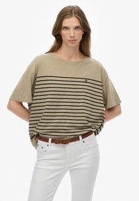 Kvinde med langt hår iført en beige oversized T-shirt med sorte striber og hvide bukser, hænderne bag ryggen, neutral baggrund.