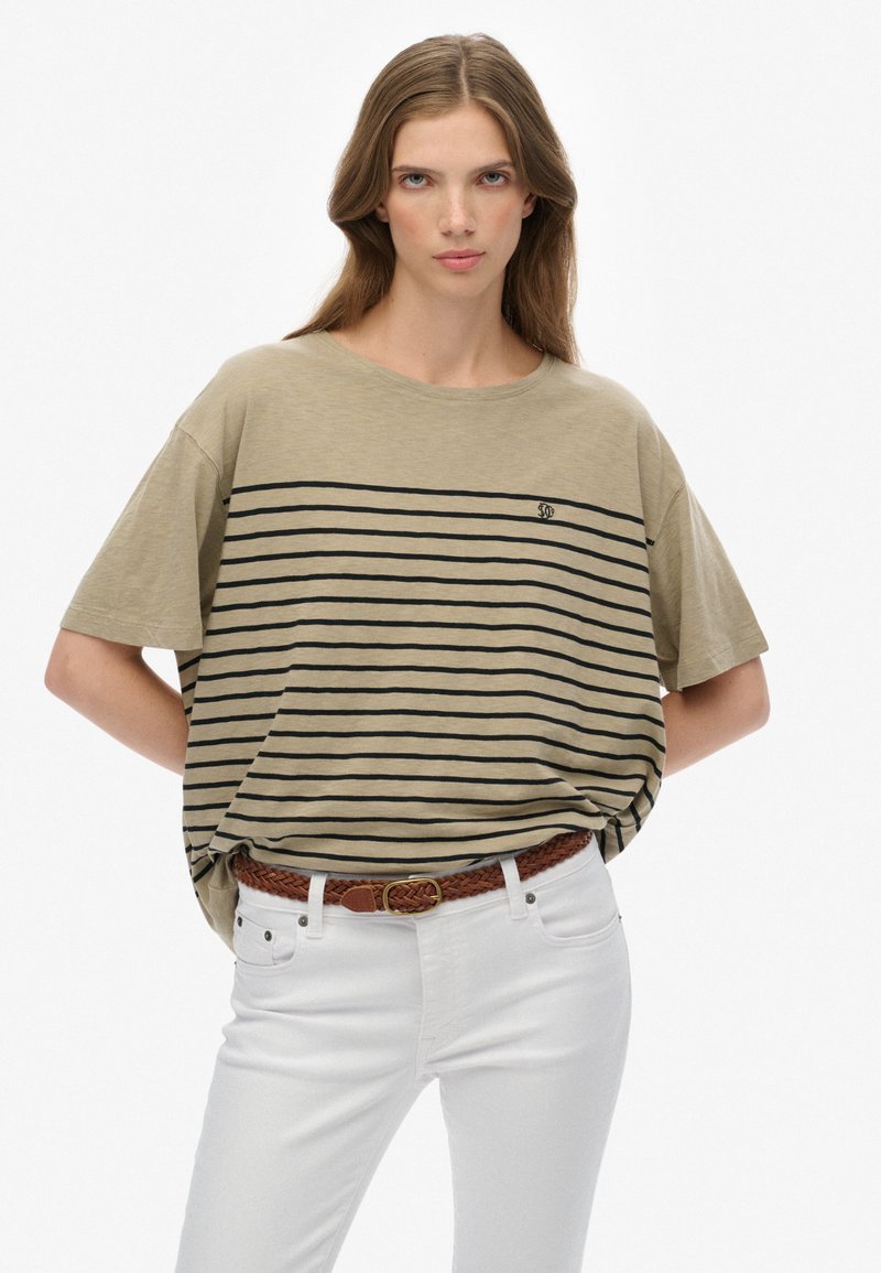 Kvinde med langt hår iført en beige oversized T-shirt med sorte striber og hvide bukser, hænderne bag ryggen, neutral baggrund.