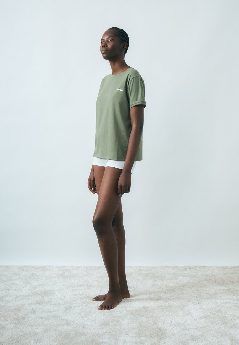 Jeune femme noire debout pieds nus sur un tapis clair, portant un t-shirt vert Hugo et un short blanc sur fond blanc uni, vue de côté.