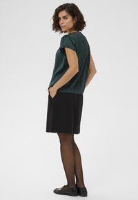 Žena s krátkymi kučeravými vlasmi, oblečená v tmavozelenej saténovej blúzke, čiernej sukni, priesvitných čiernych pančuchách a čiernych mokasínach, pózuje s rukami v vreckách.