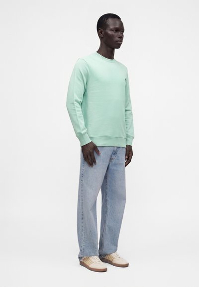 Homme debout portant un sweat-shirt vert clair, un jean bleu clair et des baskets blanches avec des détails beige et orange sur fond blanc.