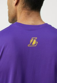 Tricou scurt, de culoare purpurie, confecționat dintr-un material moale, cu un logo galben și negru pe spate, care reprezintă o minge de baschet și litera "L".