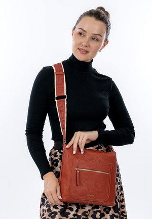 Woman in black turtleneck and leopard skirt holding textured brown leather crossbody bag with wide strap and front zipper pocket.
