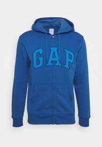 Sweat à capuche zippé bleu avec poches avant, capuche avec cordon de serrage, et grand logo "GAP" en lettres majuscules sur la poitrine.