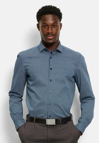 Blau gemustertes Langarmhemd mit einem Button-Down-Kragen und subtilen geometrischen Designs. Getragen mit dunklen Hosen und einem schwarzen Gürtel.