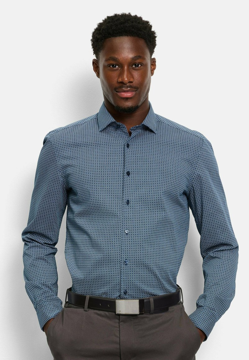 Blau gemustertes Langarmhemd mit einem Button-Down-Kragen und subtilen geometrischen Designs. Getragen mit dunklen Hosen und einem schwarzen Gürtel.
