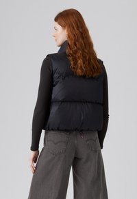 Zwarte puffervest met hoge kraag, mouwloos ontwerp, gestikte secties en een gladde textuur. Gecombineerd met grijze wijde broeken.