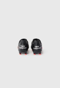 Des crampons de football noirs avec des accents rouges, présentant une surface texturée, des trous de ventilation et une marque "new balance" sur le talon.