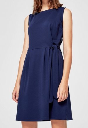 Robe sans manches bleu marine longueur genou avec un détail de nœud latéral portée par une femme debout sur un fond uni.