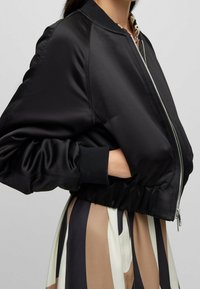 Svart bomberjacka med en slät satinfinish, ribbade muddar och midja, med en framzip och sidfickor.