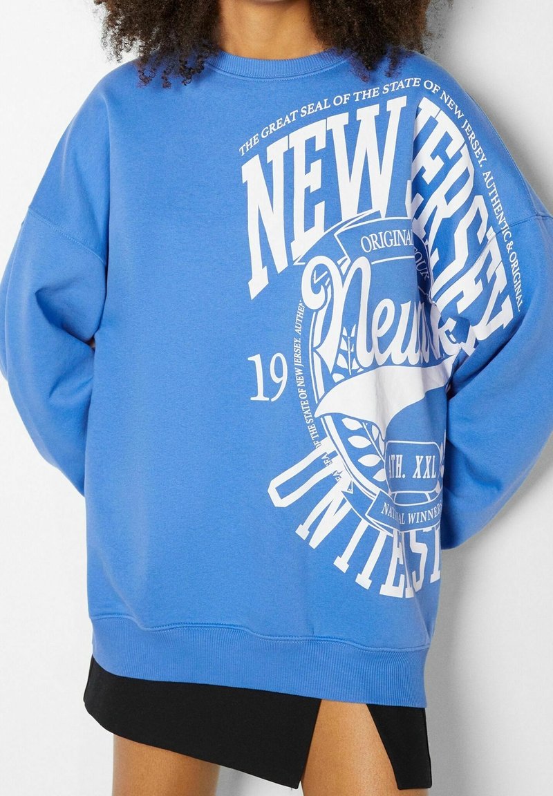 Osoba má na sobě modrý volný svetr s bílým nápisem "New Jersey University" a znakem, doplněný černou sukní s rozparkem na stehně.